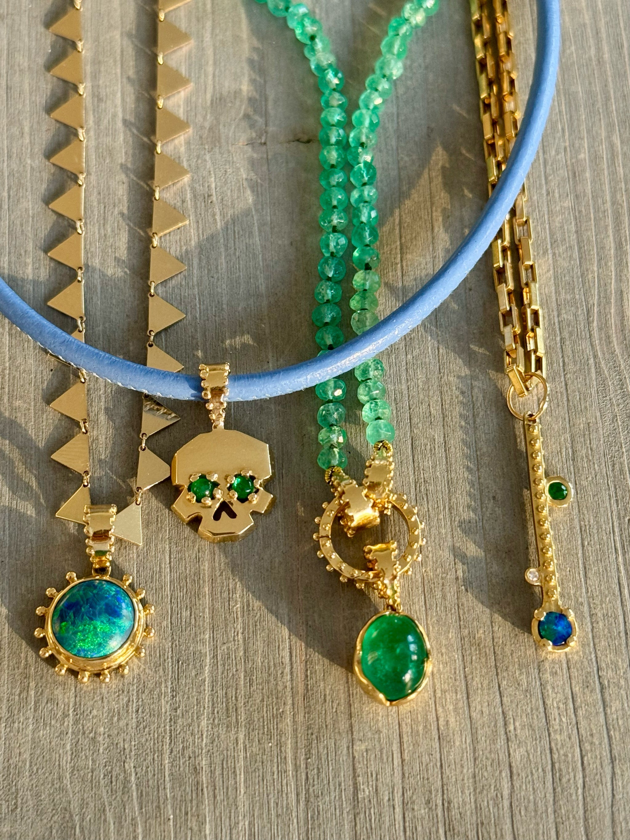 Flat lay of Delphine Leymarie Green Magic Edit with emerald, opal, turquoise, and solid gold talisman necklaces, including skull, sun, triangle, and gemstone bead layers.