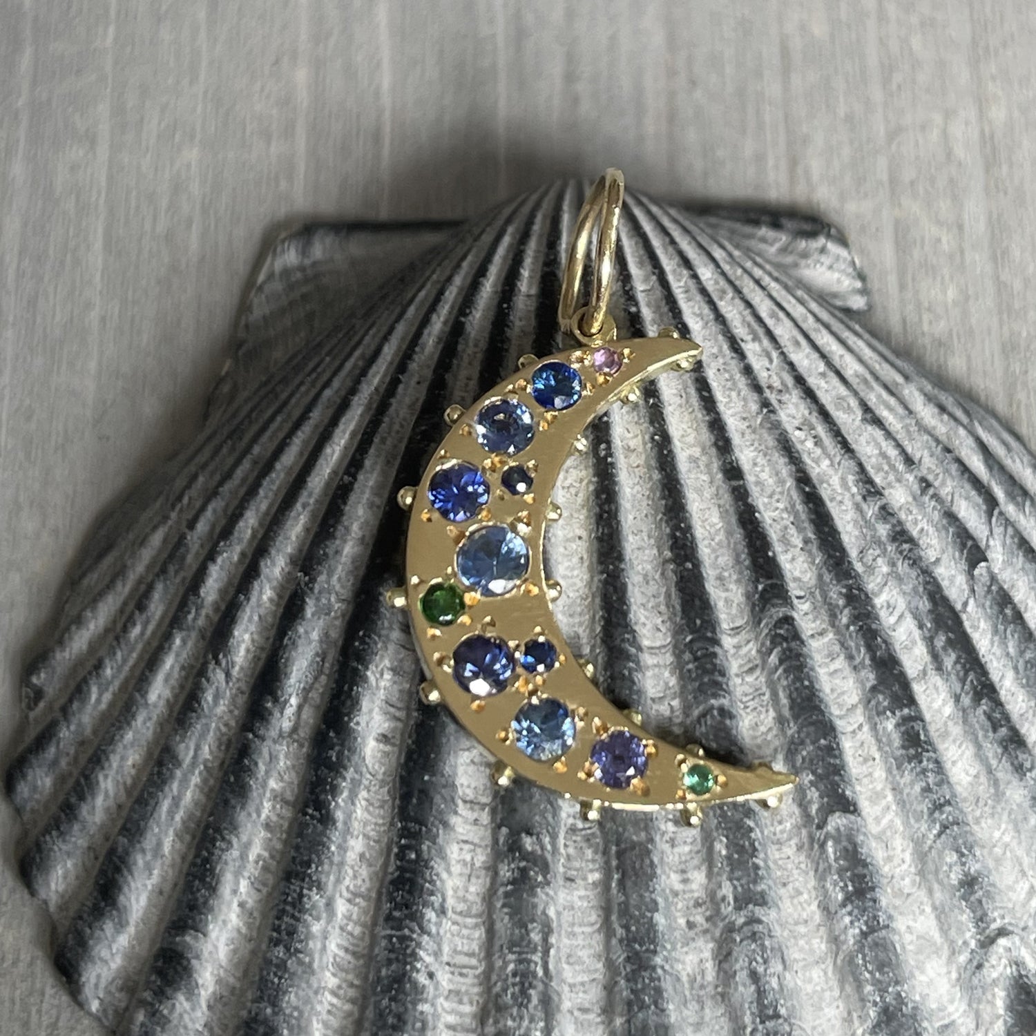 An 18k yellow recycle gold crescent moon charm with blue, purple sapphire, green tsavorite, and amethyst on a grey seashell.