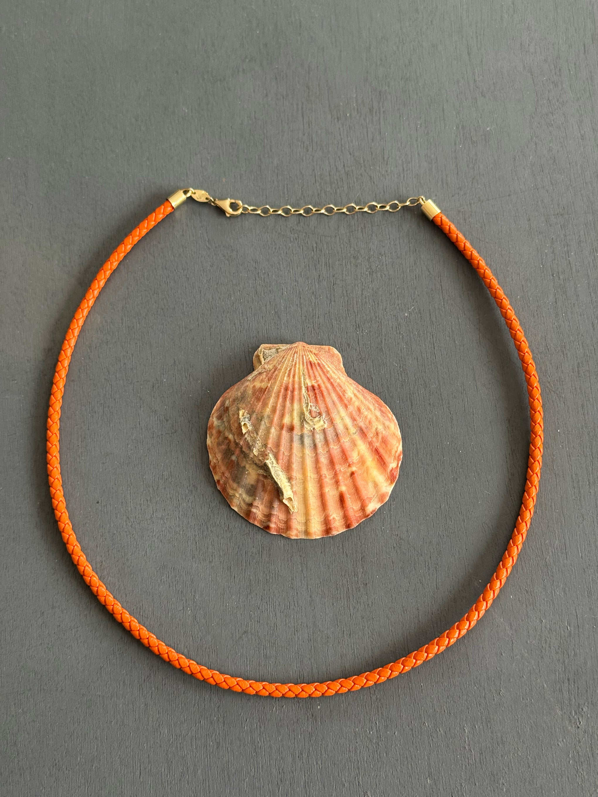 Sacred Link orange braided leather cord with solid 14k yellow gold clasp and extender, displayed on a grey background around an orange seashell.