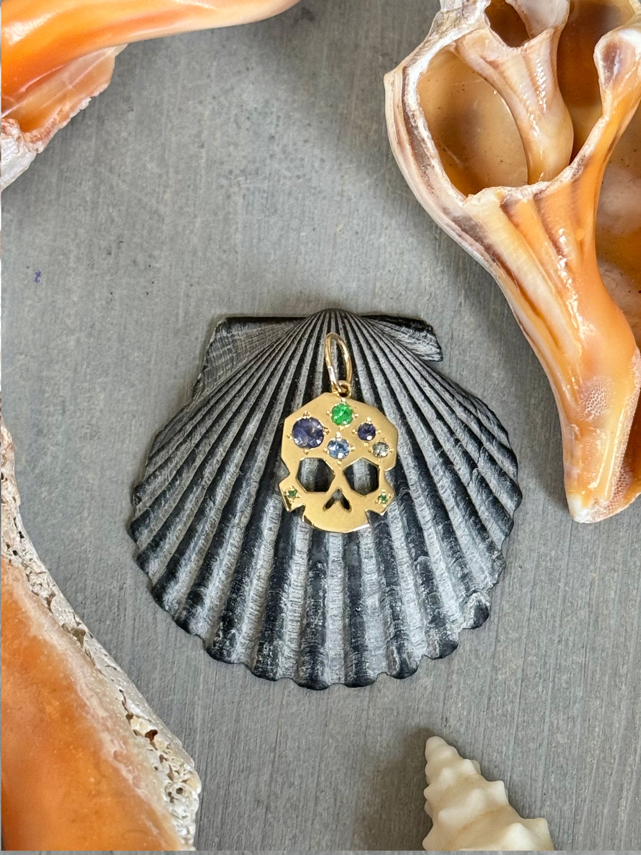 OOAK Facette Skull charm in 18k gold with blue, lavender, and green gems, styled on a dark scallop shell and surrounded by natural seashells on grey wood.