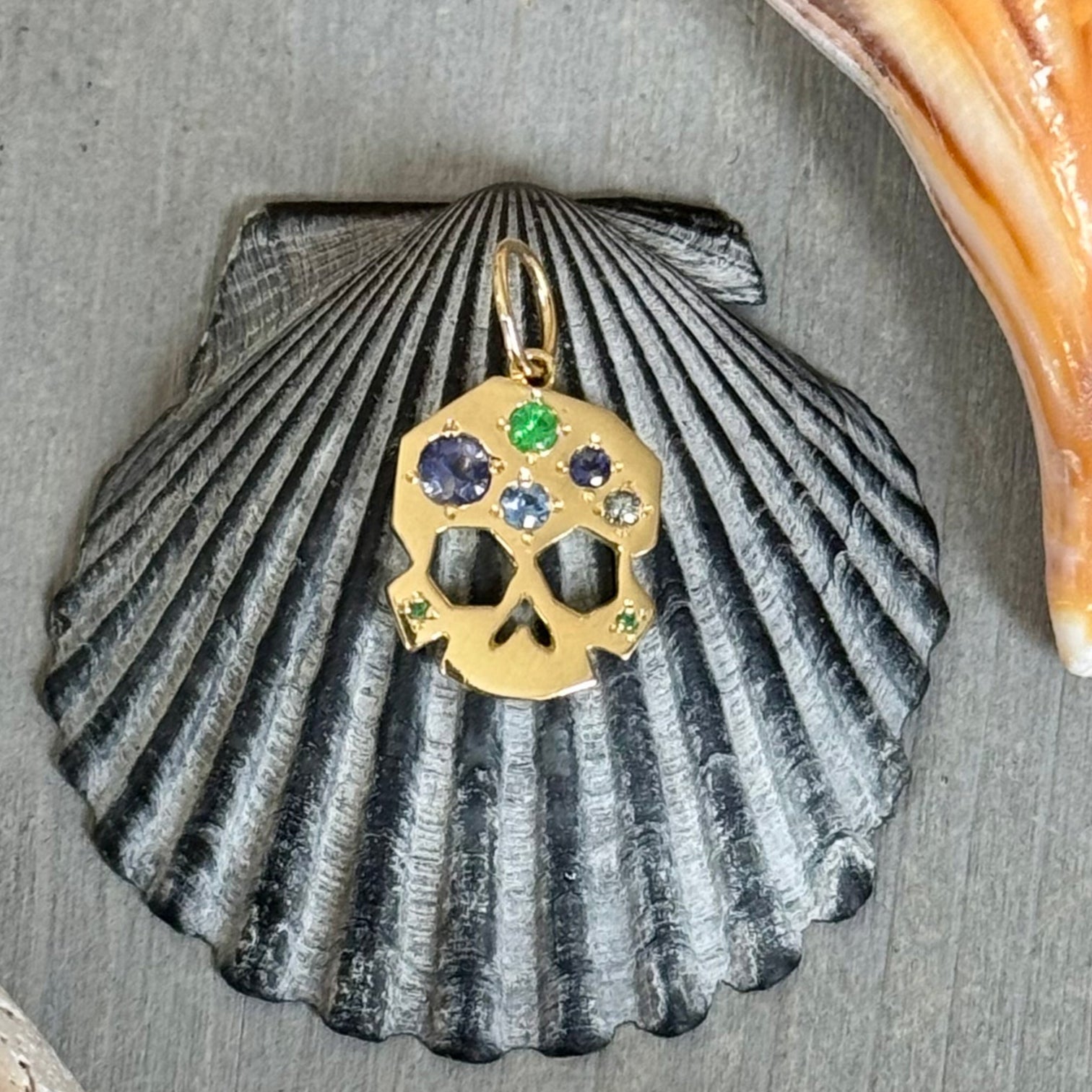 OOAK Facette Skull charm in 18k gold with blue, lavender, and green gems, styled on a dark scallop shell and surrounded by natural seashells on grey wood.