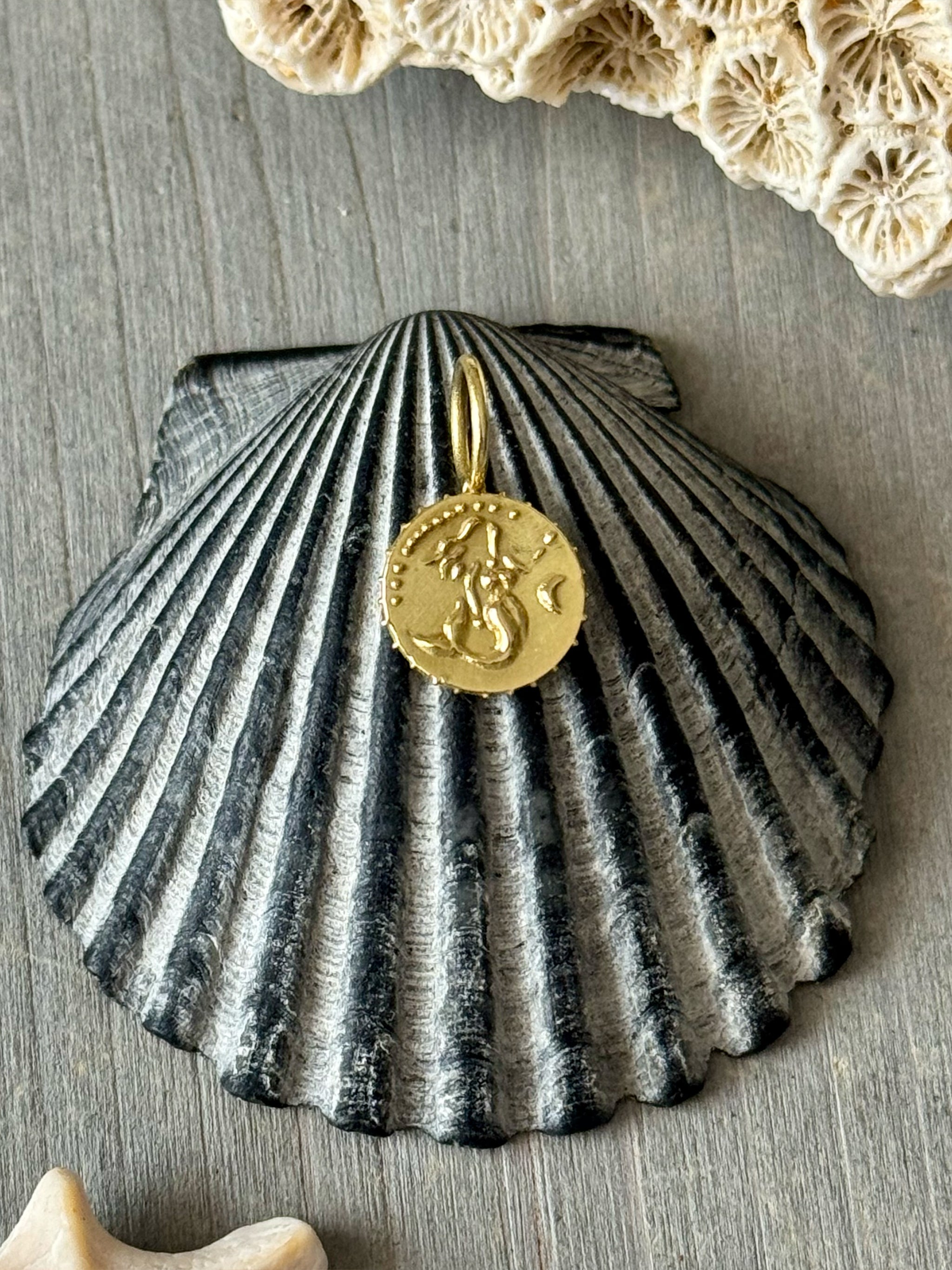 Solid 18k recycled gold tiny mermaid medallion talisman charm on a grey shell with seashells in the background