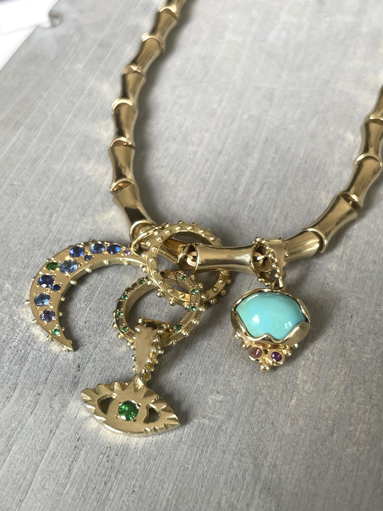 Boheme Turquoise Lotus and tsavorite Eye charm, Blue Sapphire Crescent Moon connected to the Bambou chain with 2 Clickers laying over Grey driftwood