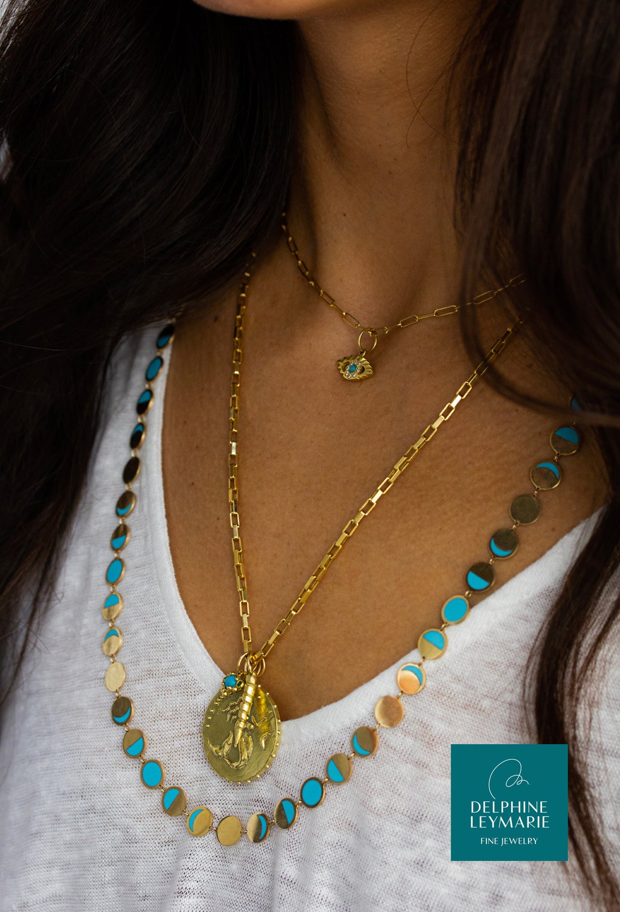 Woman wearing 18k yellow recycled gold necklaces including Unicorn horn, mermaid, lotus and eye talismans, layered with turquoise inlay Confetti Lune Moon phase chain and Delphine Leymarie logo.