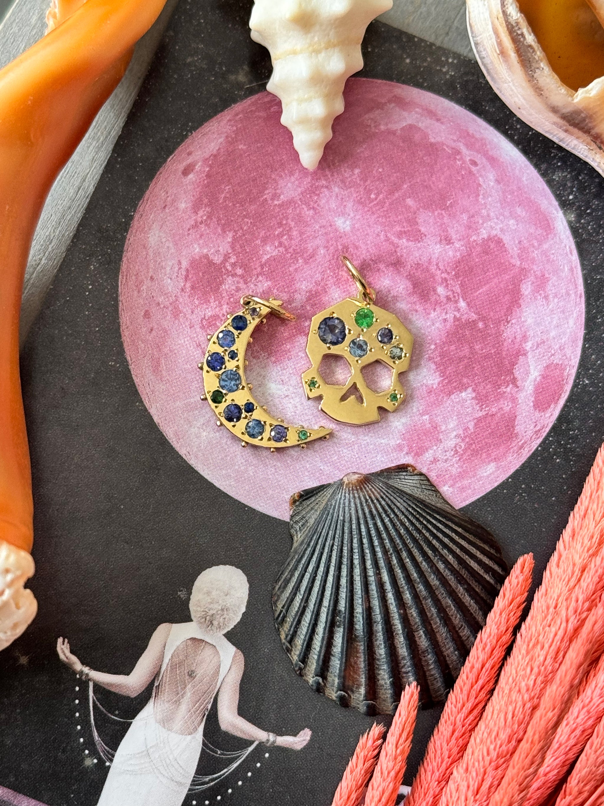 OOAK Facette Skull charm and Boheme Sapphire Crescent Moon charm in 18k gold, styled atop a pink moon tarot card with seashells and coral branches.