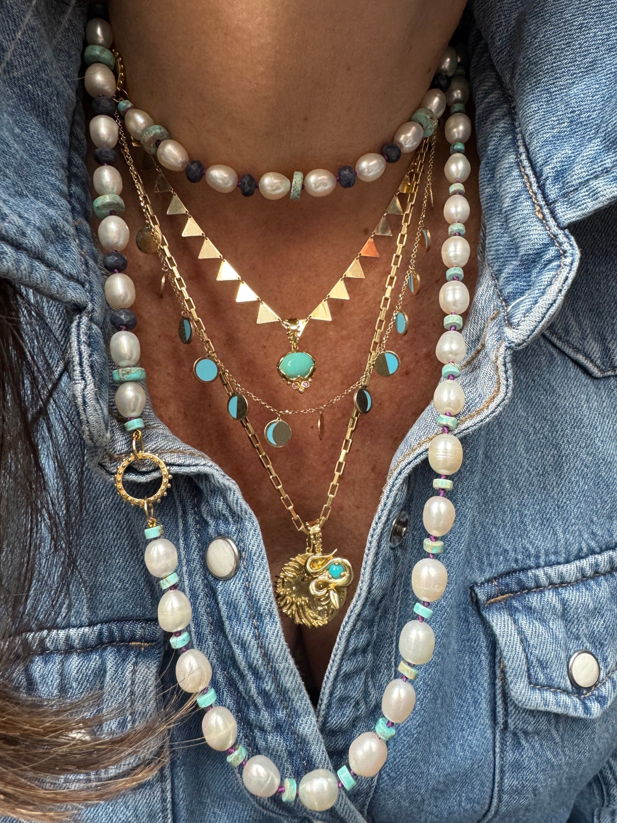 Layered turquoise and gold talisman necklaces with lion, snake, lotus, and moon symbols, styled with pearl strands over denim.
