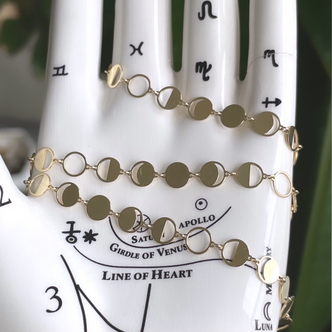 A gold chain necklace with circular cutouts featuring moon phases wrapped around a porcelain hand.