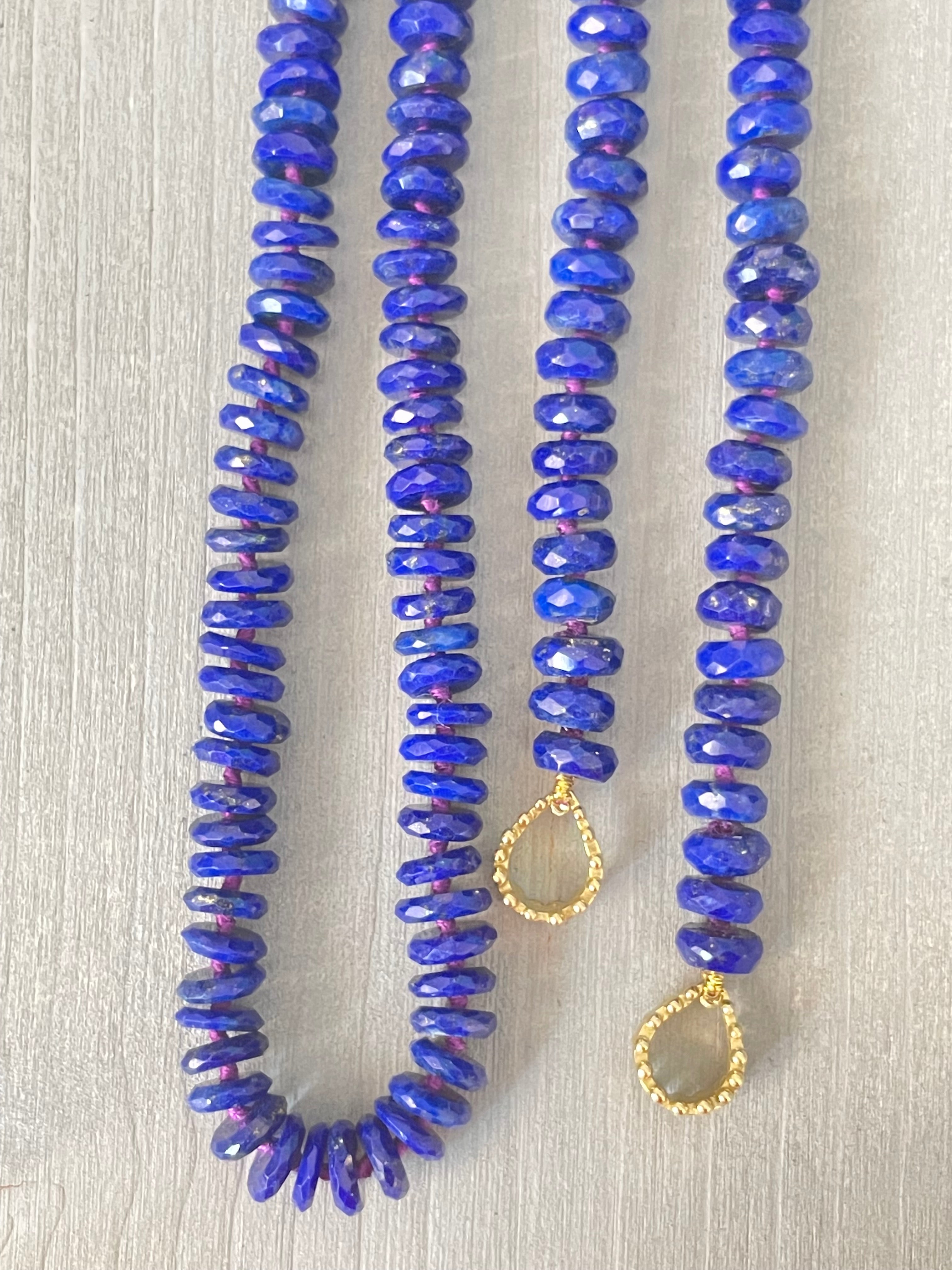 A strand of Delphine Leymarie deep blue lazuli beads with gold pyrite accents on purple silk thread with gold beaded tear drop hoops at each end, displayed on a gray driftwood.