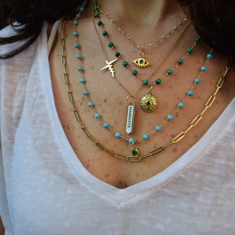 Emily wearing gold layers including Maman in white enamel, Tsavorite Urchin charm, Tsavorite Eye, Heartbeat necklace and wire-wrapped Delph chains in turquoise and malachite