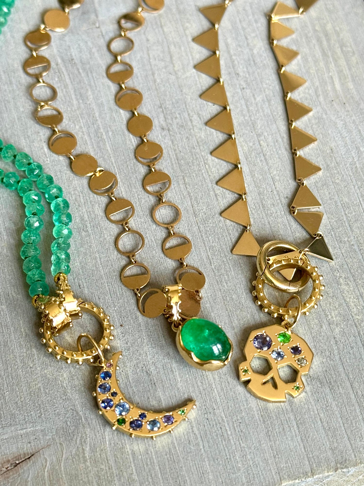 Solid gold crescent moon, skull, and emerald lotus charms on necklaces styled on grey wood—Beltane ritual jewelry flat lay.
