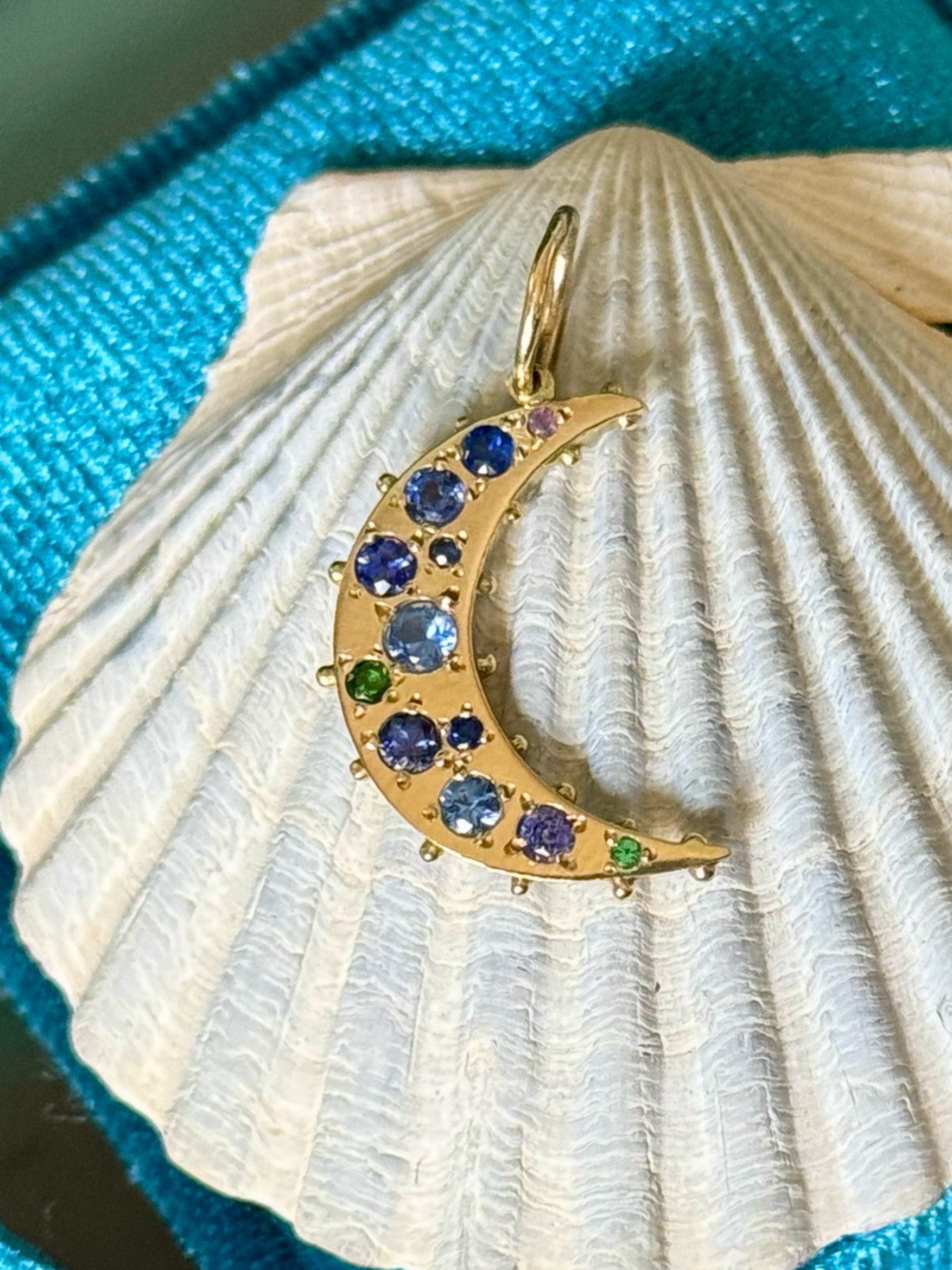 18k recycled yellow Gold crescent moon pendant with blue pruple sapphire on a seashell against a blue velvet background