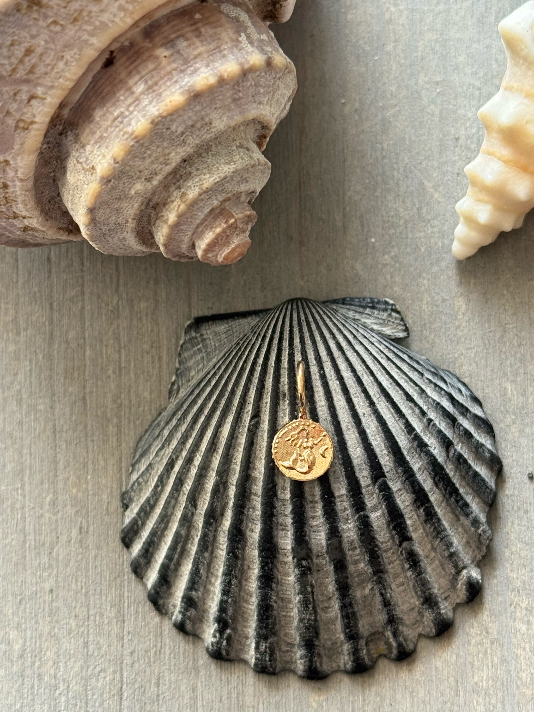 Tiny 18k gold Bébé Sirène mermaid charm resting on a dark scallop shell with natural sea shells by Delphine Leymarie