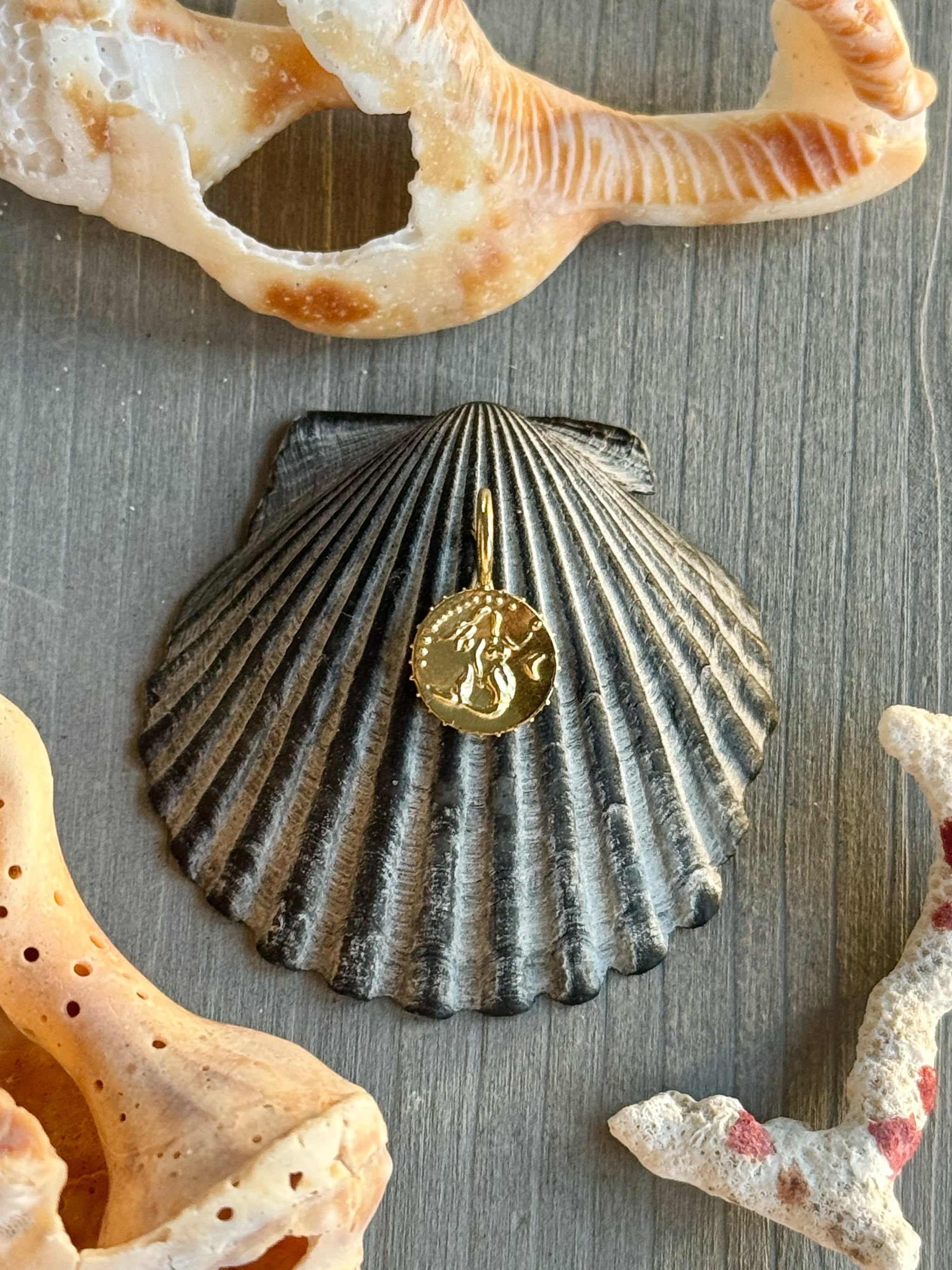 Solid 18k recycled gold tiny mermaid medallion pendant on a grey shell with seashells and coral in the background