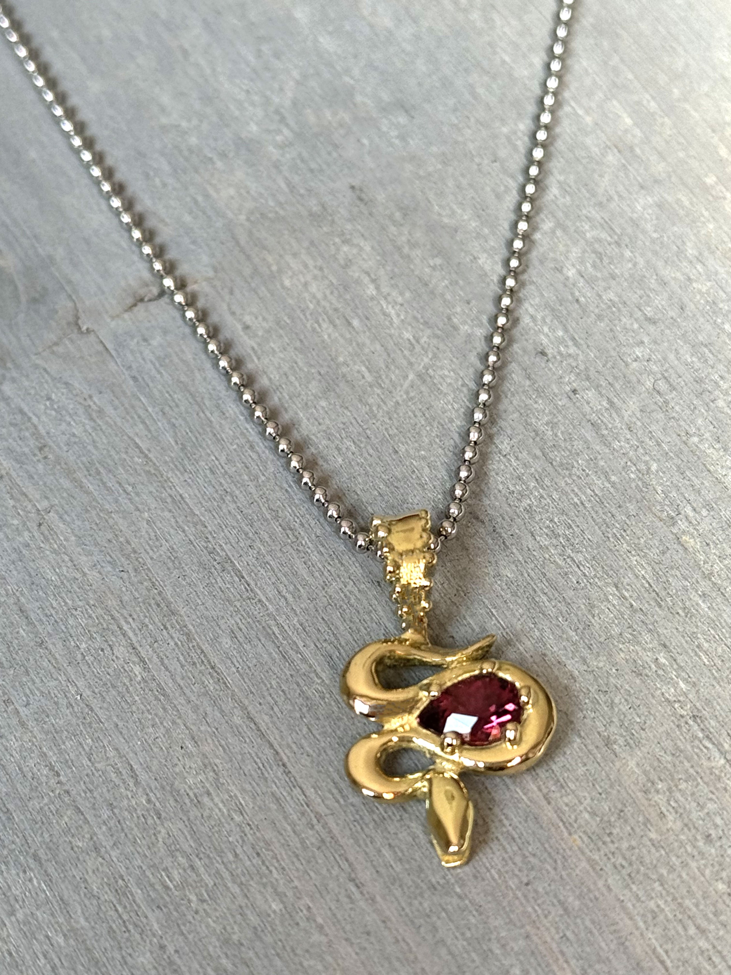 A close-up of the Bohème Serpent Charm in luminous 18 karat yellow Fairmined gold, coiled around a vivid raspberry Rhodolite Garnet. The serpent's intricate details catch the light, set against a grey wood background.