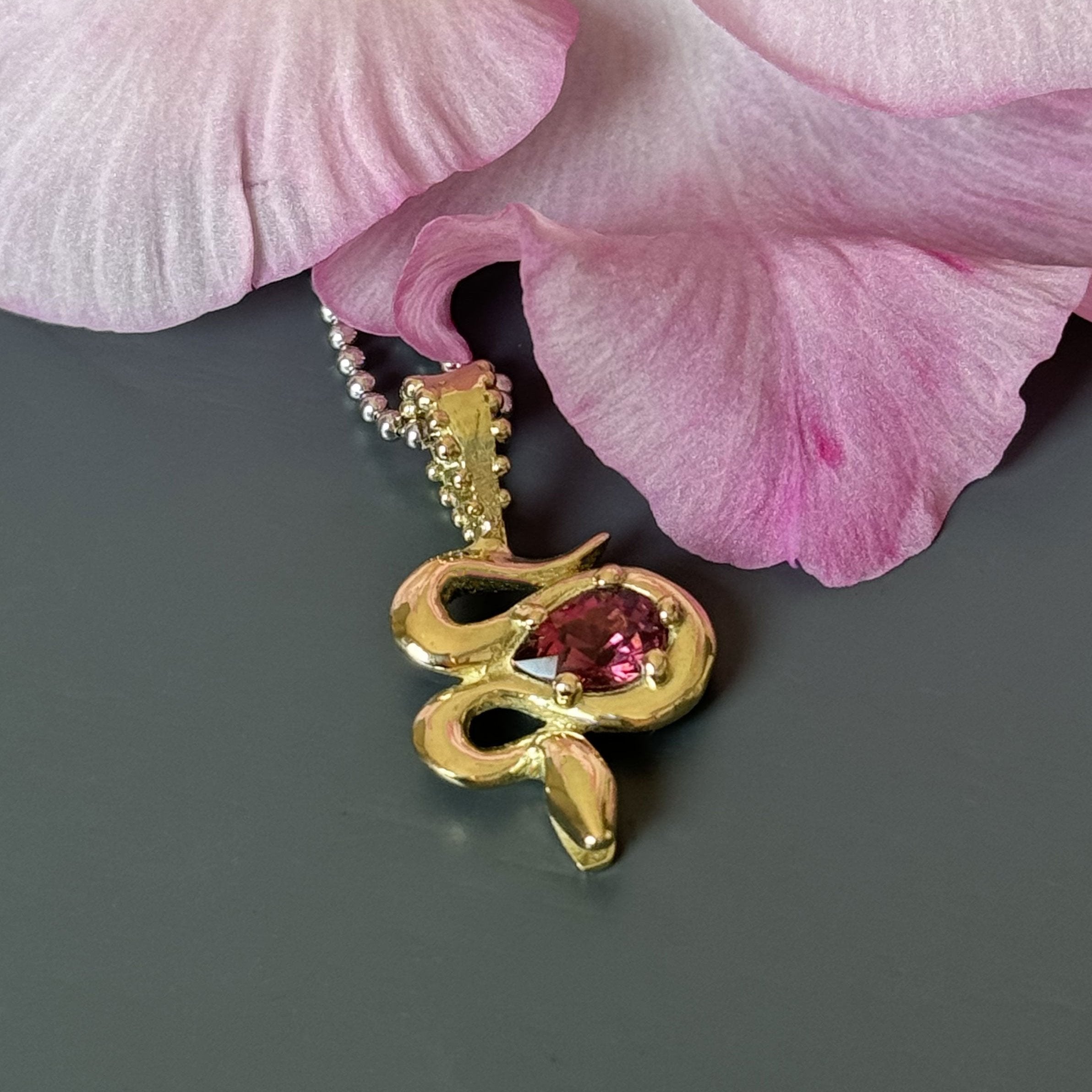 A close-up of the Bohème Serpent Charm in luminous 18 karat yellow Fairmined gold, coiled around a vivid raspberry Rhodolite Garnet. The serpent's intricate details catch the light, set against a dramatic grey background with a soft pink lily accent.