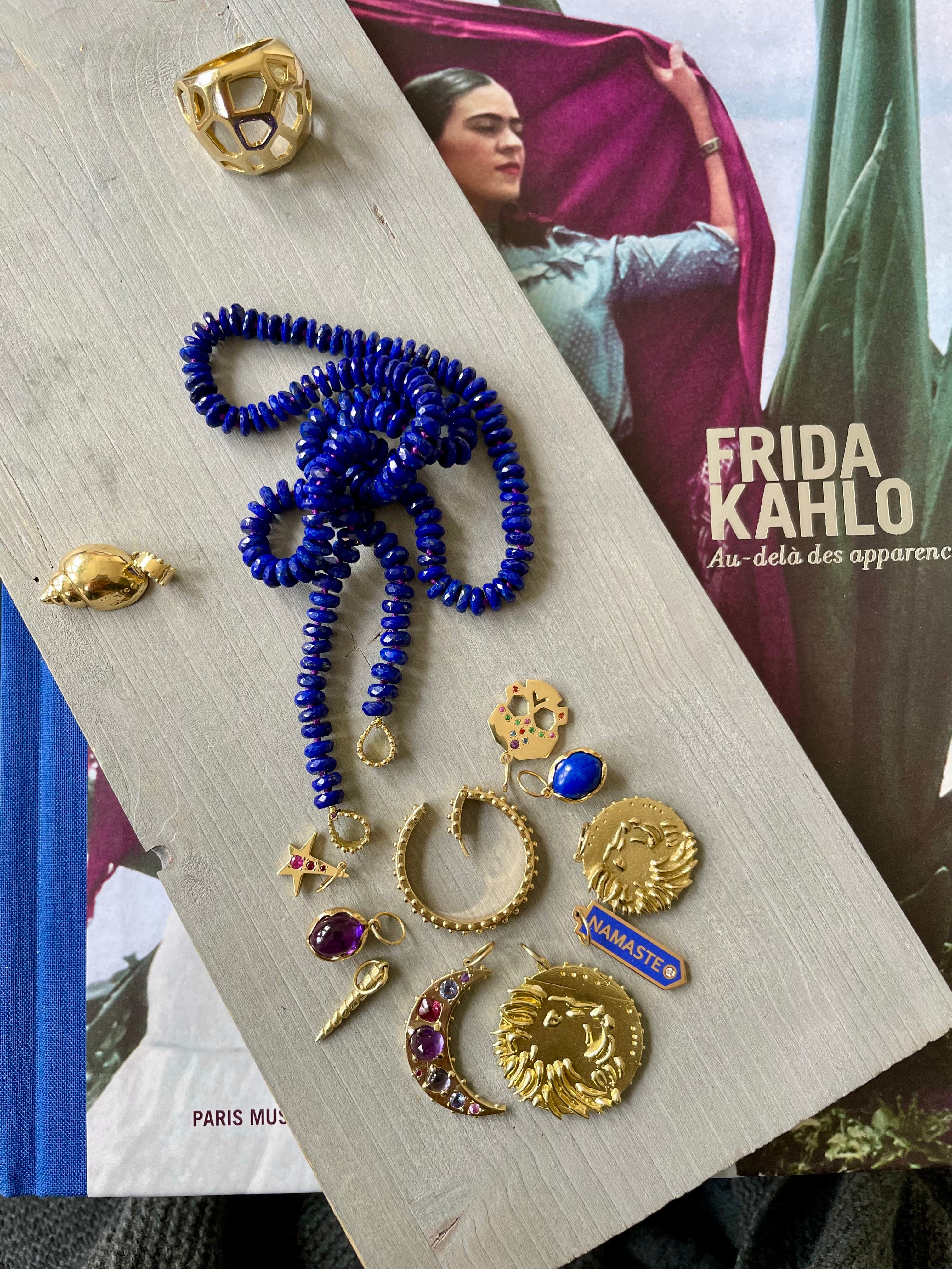 Jewelry set including a Delphine Leymarie beaded lapis necklace and gold talismans on a wooden board with a Frida Kahlo book in the background.