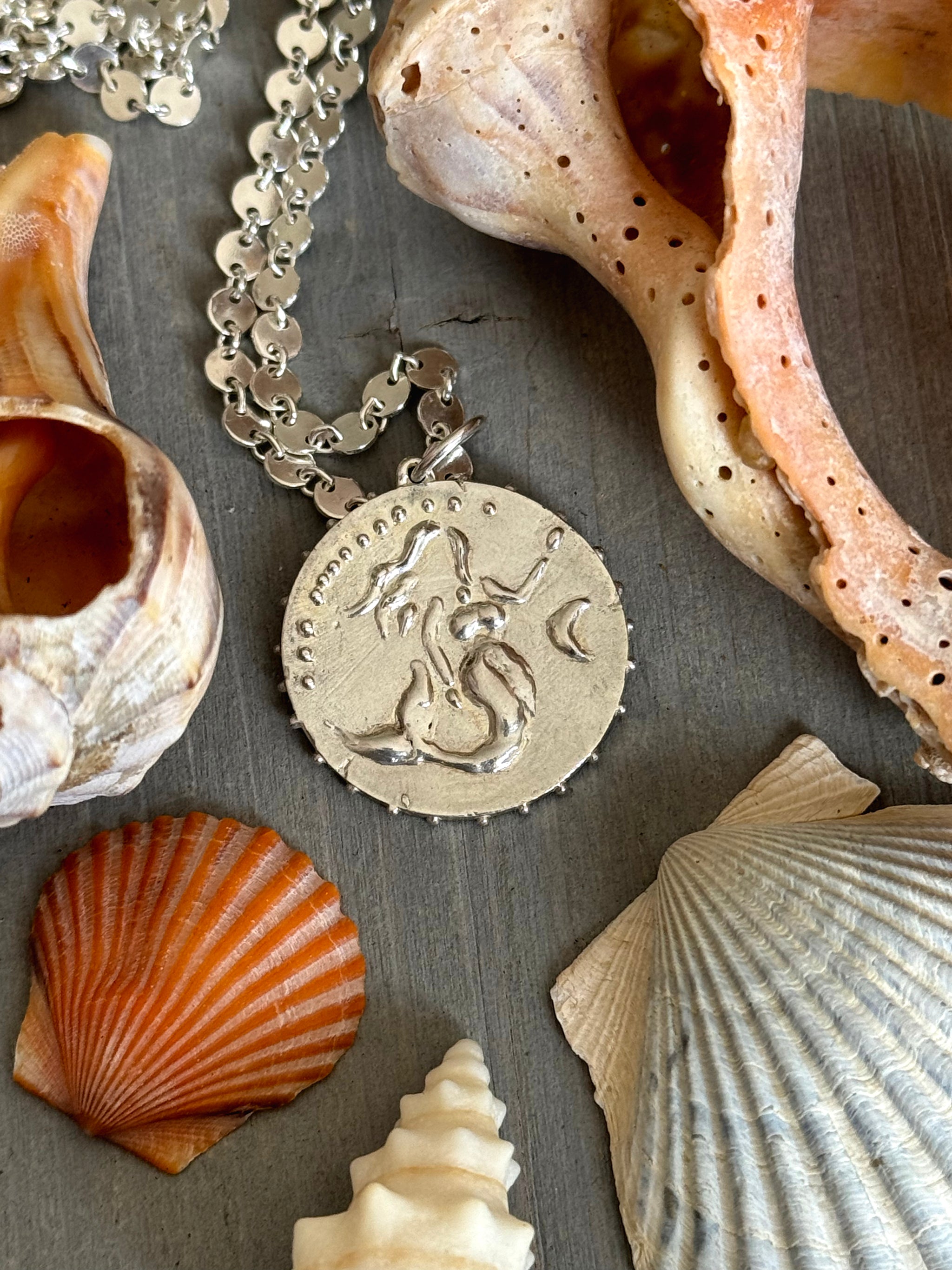 Flat lay of Delphine Leymarie Boheme Grande Sirene Medallion mermaid on Disco chain in Silver over Grey Wood surrounded by natural seashells.