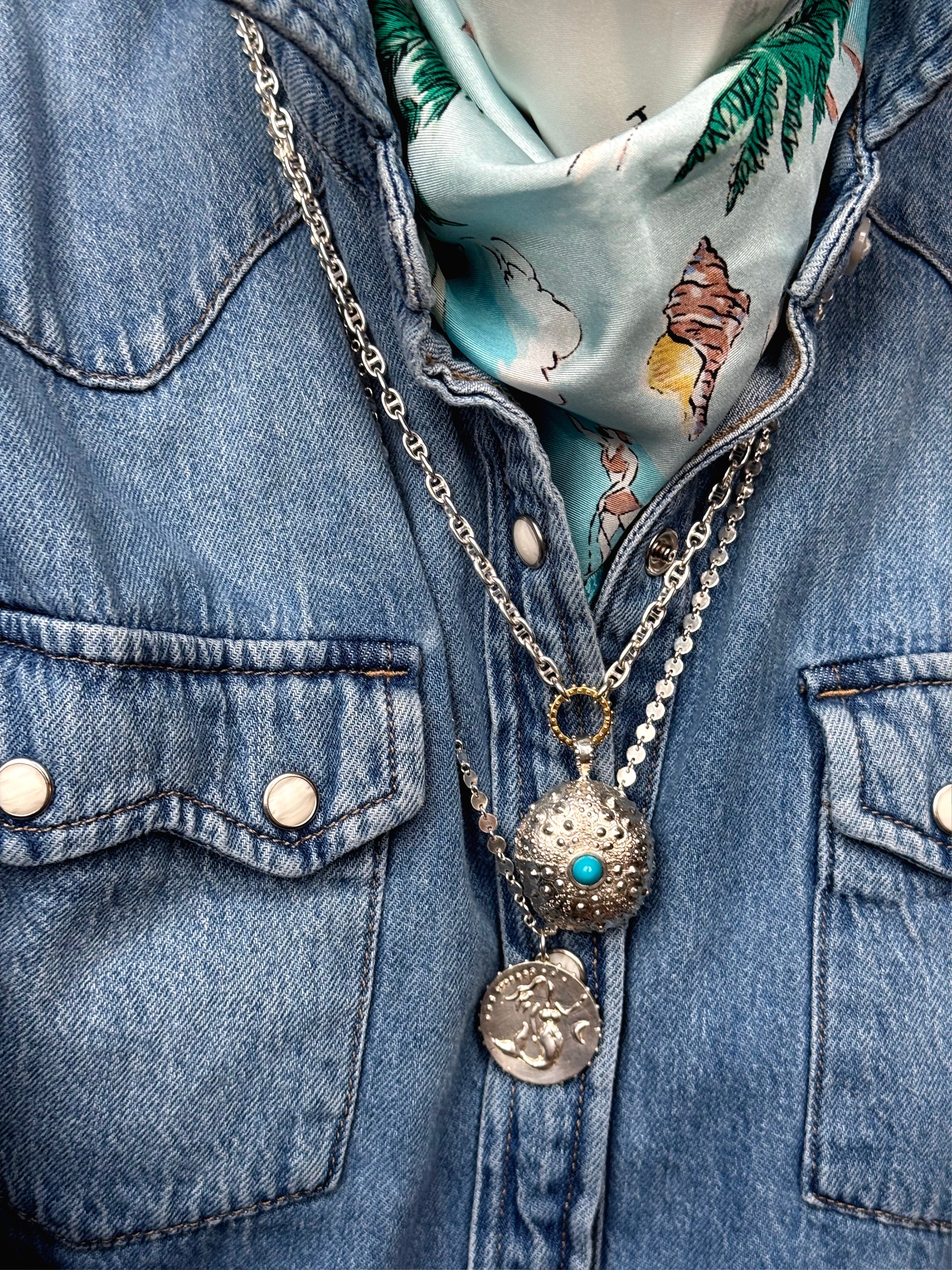 Layered silver necklaces over a denim shirt and silk scarf, featuring a silver sea urchin pendant with turquoise center and a mermaid medallion by Delphine Leymarie