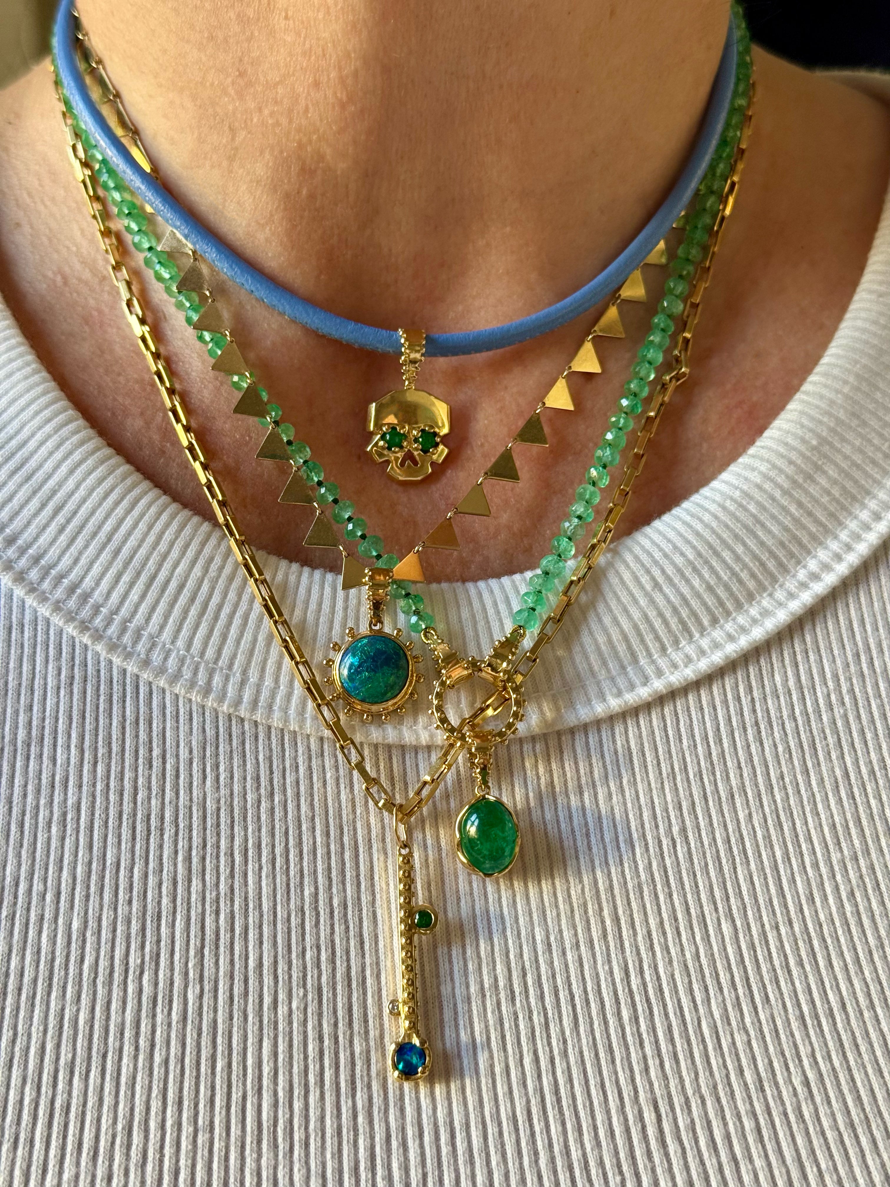 Layered Delphine Leymarie 18k solid gold talisman necklaces with emerald skull charm, opal sun charm, emerald beads, blue cord choker, and triangle chain.